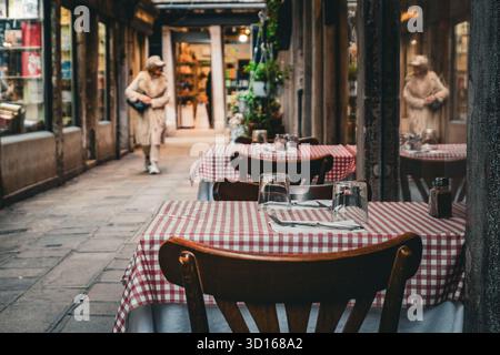 Tavoli vuoti all'aperto del ristorante con tovaglie a scacchi in uno stretto vicolo veneziano, Venezia, Italia, in una giornata nuvolosa Foto Stock