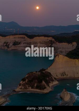 Scatto aereo verticale di una luna piena luminosa che si innalza sulle spettacolari scogliere bianche di Capo Drastis, Corfù. I fondali marini e la costa sono illuminati dal twili Foto Stock