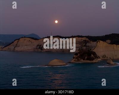 Una luminosa luna piena sorge in un cielo viola crepuscolo sopra le spettacolari scogliere bianche e le montagne marine di Capo Drastis, Corfù, con il suo riflesso sul cal Foto Stock