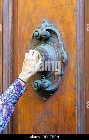 Primo piano di una mano che stringe un pomello della porta vecchio stile montato su una contropiastra in ferro ornato, fissata a una porta in legno resistente agli agenti atmosferici. L'immagine trasmette il mister Foto Stock
