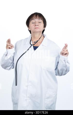 Medico donna che indossa un camice da laboratorio e uno stetoscopio, in piedi con gli occhi chiusi e le dita incrociate su entrambe le mani Foto Stock