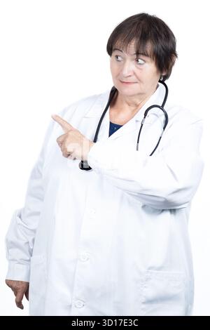 Medico anziano in camice da laboratorio bianco e punti stetoscopio lasciati con l'indice, espressione seria che trasmette avvertenze, istruzioni Foto Stock
