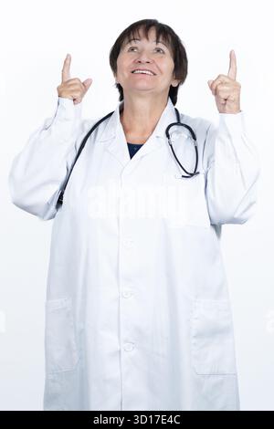 Donna matura che indossa un camice da laboratorio bianco e uno stetoscopio, che punta felicemente entrambe le dita indice verso l'alto, guarda verso l'alto e sorride Foto Stock
