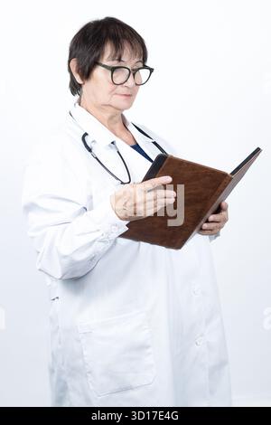 Medico donna maturo in camice da laboratorio e stetoscopio, con occhiali, in piedi e studiando un libro di registrazione del paziente, specializzato e professionale Foto Stock