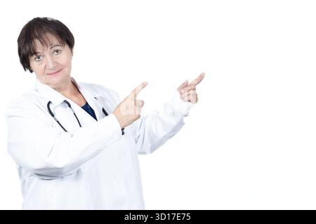 Medico esperto che indossa un camice da laboratorio e uno stetoscopio, sorridendo mentre punta due dita verso uno spazio vuoto su uno sfondo bianco, suggerendo Foto Stock