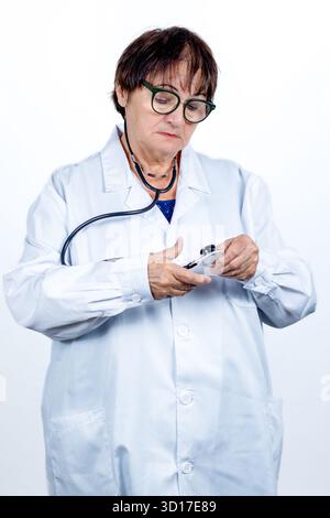 Medico donna anziana che indossa un camice da laboratorio bianco e occhiali, tenendo e regolando il suo stetoscopio, simboleggiando l'assistenza sanitaria, la professionalità Foto Stock