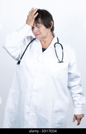 Donna anziana che indossa un camice da laboratorio bianco e uno stetoscopio che soffre di grave mal di testa o emicrania, sentendosi sfinita Foto Stock