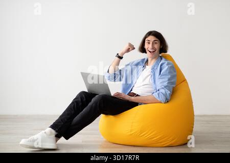 Giovane uomo che lavora su un laptop seduto su una sedia con borsa in fagioli gialli con espressione gioiosa Foto Stock
