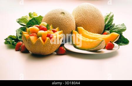 Una composizione decorativa di frutta con due cantalopi interi, due fette di cantalope su un piatto e un cantalopo scolpito in un cesto pieno di Foto Stock
