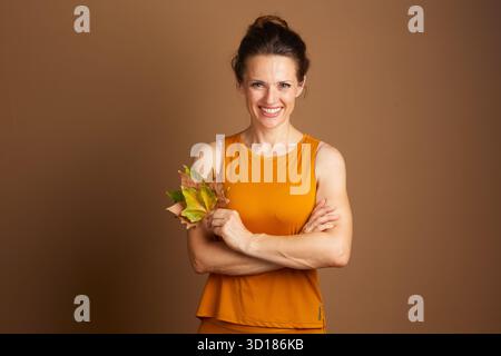 Una donna adulta sorridente con capelli scuri, con canotta color senape, si stende con le braccia incrociate, tenendo le foglie autunnali su un dorso marrone pieno Foto Stock