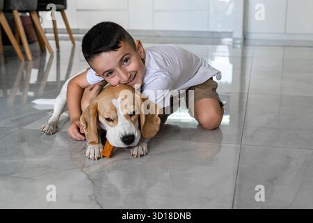 Un ragazzo allegro abbraccia il suo beagle su un pavimento lucente, mentre il cane si concentra sulla masticazione di un gustoso regalo. Questa scena cattura un momento caldo tra un animaletto giocoso e un bambino felice. Foto Stock