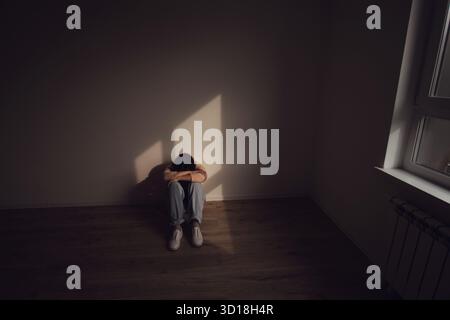 Una donna siede da sola sul pavimento in una stanza poco illuminata, abbracciando le ginocchia, esprimendo tristezza, isolamento e angoscia emotiva. Foto Stock