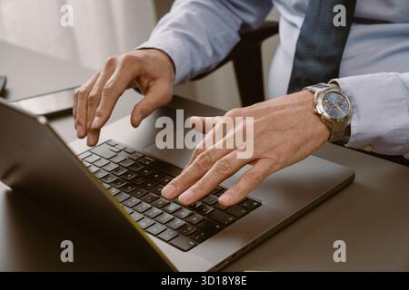 Un uomo è concentrato sul suo notebook, scrivendo con attenzione mentre è seduto alla scrivania ordinata. Uno smartphone e un blocco note si trovano nelle vicinanze, a indicare un ambiente di lavoro occupato Foto Stock