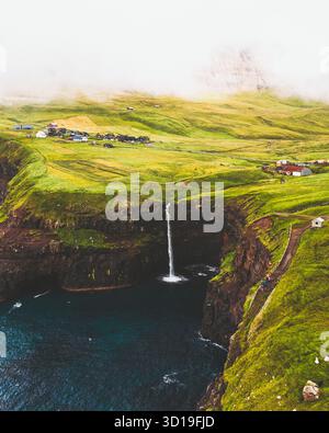 La vista aerea della cascata di Múlafossur si tuffa nel mare blu scuro, circondato da scogliere verdeggianti e dal pittoresco villaggio di Gasadalur, Vágar, Isole Faroe. Foto Stock