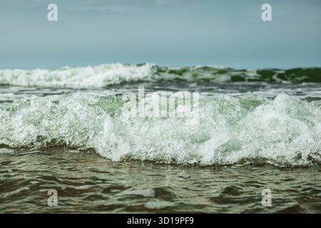 Piccole onde di marea schiumose che raggiungono la riva, primo piano, angolo basso, punto di vista del livello dell'acqua, nuvoloso giorno d'estate, nessuna gente Foto Stock