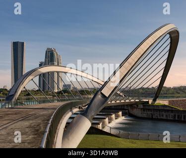 Ponte pedonale sul lago a Taman seri Empangan, Putrajaya, Malesia. Moderno design ad arco su cascate, con grattacieli contemporanei visibili sotto un cielo blu limpido. Foto Stock