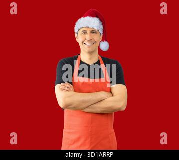 Foto di studio di un uomo ispanico allegro uomo positivo sorridente con le braccia incrociate che esprimono emozioni positive mentre indossa un cappello di Natale aga Foto Stock