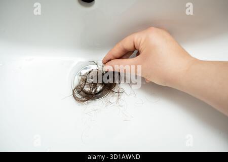 Tira a mano un panino di capelli sporchi femminili dal foro di scarico del bagno. Scarico fai-da-te intasamento pulizia scarico doccia perdita capelli stress, beriberi, alopecia. Ostruzione wa Foto Stock