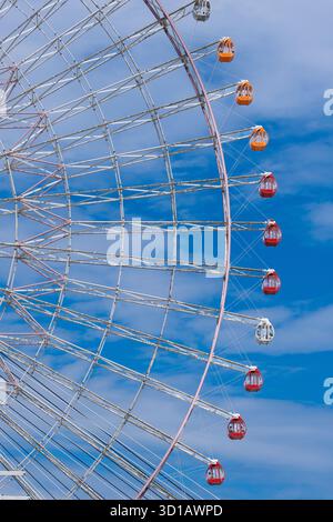 Una grande ruota panoramica con gondole rosse, arancioni e bianche vibranti poste su un cielo blu limpido con nuvole sparse. Foto Stock
