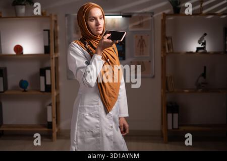Concetto di intelligenza artificiale in medicina. Medico musulmano in hijab comando ad assistente ai, dettare il messaggio audio utilizzando lo smartphone lavorando su vicino Foto Stock