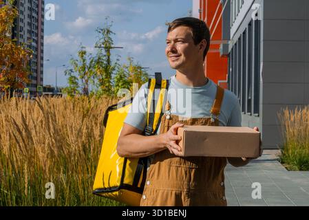 Giovane che lavora come corriere, indossa una borsa gialla e tiene in mano una scatola di cartone marrone, fornendo servizi di spedizione efficienti in un ambiente urbano Foto Stock