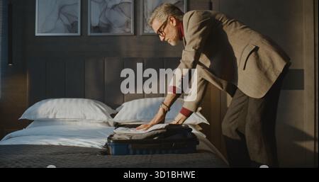 L'elegante uomo dai capelli grigi confeziona meticolosamente i vestiti in una piccola valigetta blu. Un uomo che ha lasciato l'hotel in fretta per prendere il volo. Concetto di viaggio, routine del viaggiatore, viaggio d'affari. Colpo statico. Foto Stock