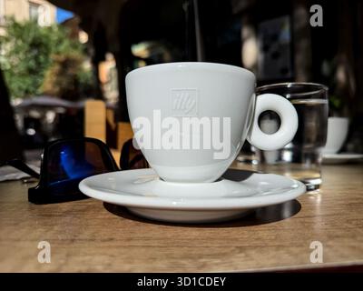 Primo piano di una tazza di espresso Illy su un tavolino da caffè all'aperto in legno con occhiali da sole e un bicchiere d'acqua Foto Stock