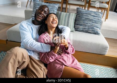 Giovani coppie diverse che sorridono gioiosamente insieme nel loro luminoso soggiorno, indossando abiti casual Foto Stock
