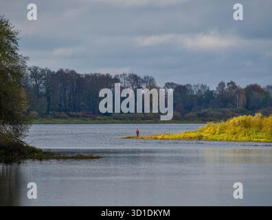 Ampio paesaggio fluviale autunnale - vista serena e grandangolare di un ampio fiume in autunno sotto un cielo nuvoloso e un mantello giallo brillante con un pescatore singolo Foto Stock