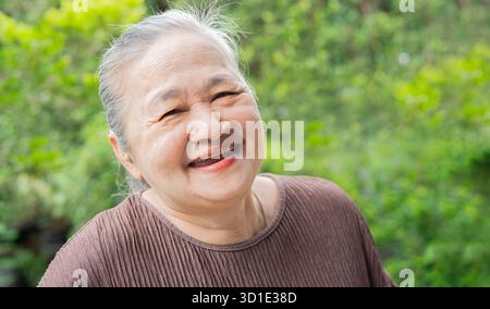 Una donna asiatica gioiosa e anziana nel giardino di casa viene catturata in un allegro ritratto, che irradia felicità con il suo sorriso caldo Foto Stock