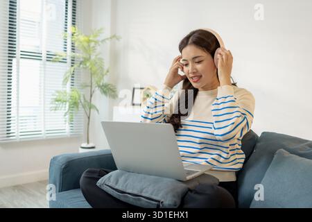 Una giovane donna che indossa le cuffie si siede comodamente sul divano, sorridendo mentre lavora al laptop. Sembra concentrata e felice, rappresentando l'architettura moderna Foto Stock