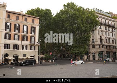 Ampia piazza lastricata dominata da grandi alberi, caratterizzata da edifici romani classici e da persone che camminano lungo la piazza vicino a un ingresso della metropolitana. Foto Stock