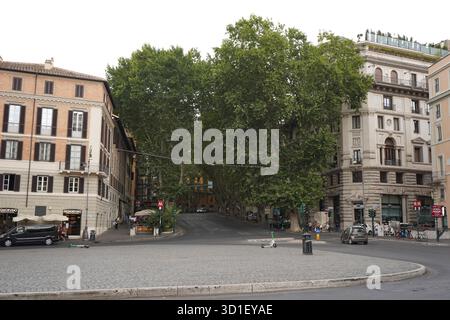 Ampia piazza lastricata dominata da grandi alberi, caratterizzata da edifici romani classici e da persone che camminano lungo la piazza vicino a un ingresso della metropolitana. Foto Stock