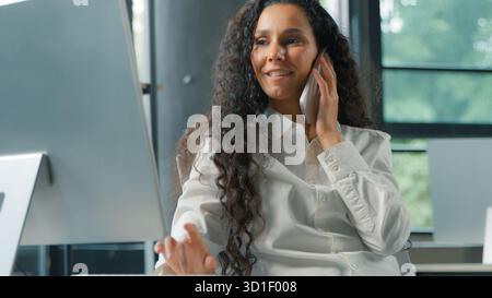 Donna d'affari ispanica sorridente che parla telefono alla ricerca di un computer donna d'affari donna donna donna datore di lavoro multitasking in ufficio lavoro professionale con Foto Stock