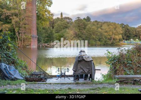 Vista posteriore del pescatore/pescatore isolato seduto con canna e attrezzatura/attrezzatura al bordo di una piscina del Regno Unito, camice sulle spalle, pesca in una fresca giornata autunnale. Foto Stock