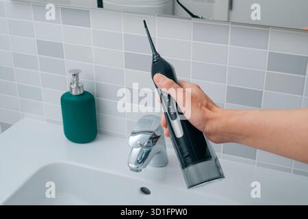 un uomo che tiene un irrigatore orale nero sopra il lavandino, in un bagno luminoso con piastrelle bianche e un dispenser di sapone verde sul bancone Foto Stock
