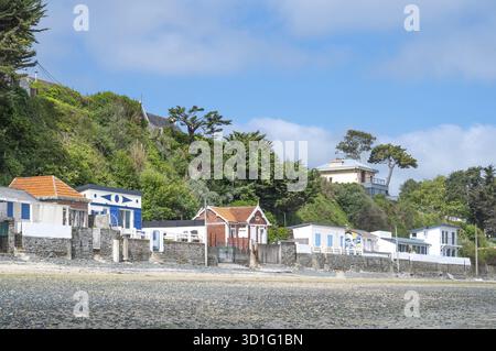 Una serie di colorate e storiche cabine sulla spiaggia con pareti di pietra naturale fiancheggiano la spiaggia rocciosa ai piedi di un ripido pendio, su cui le ville sono incastonate Foto Stock