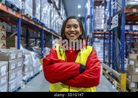 Giovane donna sorridente in piedi con le braccia incrociate in un magazzino frigorifero illuminato. Indossare indumenti protettivi rossi e gialli di sicurezza. Rappresenta e. Foto Stock