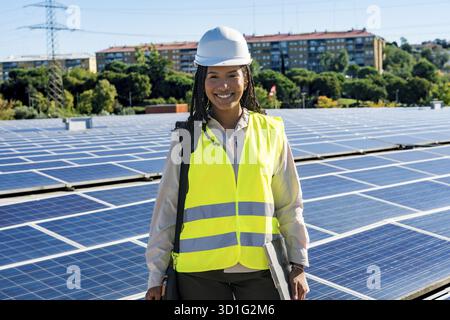 Ingegnere donna sorridente in piedi su un tetto pieno di pannelli solari, indossando un elmetto e un giubbotto di sicurezza, che rappresenta energia verde, sostenibilità e. Foto Stock
