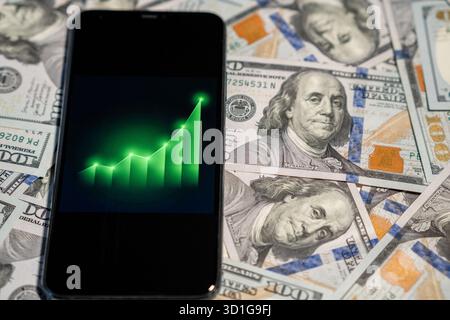 Uno smartphone mostra un grafico in aumento che simboleggia la crescita finanziaria, posizionato sullo sfondo di banconote in dollari. Foto Stock