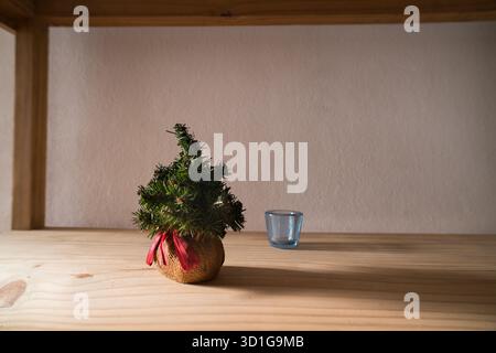 Un affascinante albero di Natale in miniatura, incastonato in una base di burlap con un nastro rosso, si erge su una mensola in legno di colore chiaro. Accanto c'è un piccolo bicchiere vuoto Foto Stock