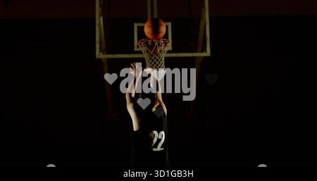 Raggiungere l'atleta che indossa la maglia numero 22 per il tiro al basket nella palestra scura, con canestro illuminato e rete Foto Stock