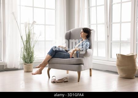 Bella signora di mezza età spensierata riposa a piedi nudi in una poltrona all'interno Foto Stock