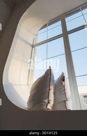 Due morbidi cuscini poggiano comodamente su un ampio davanzale della finestra, direttamente di fronte ad una grande e invitante finestra che lascia entrare il sole Foto Stock