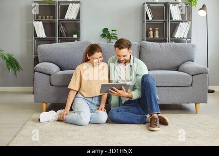Coppia felice che usa un tablet, donna allegra e uomo seduti sul pavimento del soggiorno insieme a casa Foto Stock