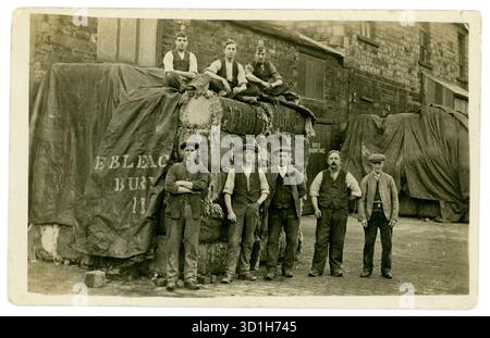 Cartolina originale dei primi anni '1900 di uomini della classe operaia in zoccoli, in piedi accanto a una pila di balle di fibra di cotone grezzo che sono coperte da sacchi assiani. Stampato su un sacco è E. Bleach Bury. Ciò potrebbe significare che le balle di cotone erano state o stavano per essere sottoposte al processo di sbiancamento prima della filatura. C'erano diverse compagnie di sbiancamento a Bury. Bury era un importante centro per la produzione tessile del cotone a causa del suo accesso all'energia idrica e, in seguito, ai motori a vapore. Dietro gli uomini c'è un edificio industriale, una fabbrica tessile di cotone o un impianto di sbiancamento. Bury, Lancashire, Regno Unito, 1912 circa. Foto Stock