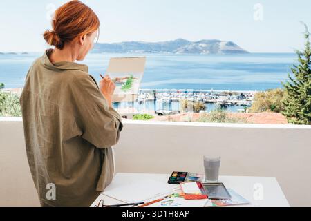 Pittura ad acquerello di donna che dipinge la vista del porto con acquerelli sulla soleggiata terrazza inondata dalla calda luce del mattino, affacciata sugli yacht e sul porticciolo di Kas Foto Stock