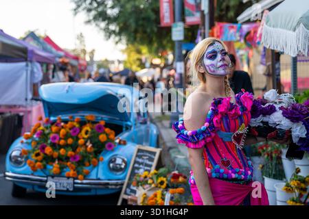 Una donna vestita da la Catrina si erge accanto a un'auto d'epoca decorata con calendule durante le celebrazioni del Día de los Muertos. Migliaia di persone si sono riunite nel quartiere culturale Barrio Logan di San Diego per onorare Día de los Muertos, una tradizione messicana che celebra la vita dei defunti cari. L'evento ha caratterizzato altari colorati, musica dal vivo e cibo di strada. Foto Stock