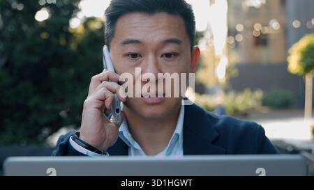 Serio uomo d'affari asiatico parlare telefono cellulare portatile utilizzando un computer di aspetto uomo d'affari coreano discutere lavoro in città parlare parlare parlare smartphone Foto Stock