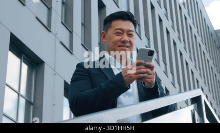 Uomo d'affari asiatico di mezza età sorridente scrivendo un telefono cellulare in città vicino all'edificio degli uffici felice uomo d'affari giapponese che usa lo smartphone in piedi sulle scale Foto Stock